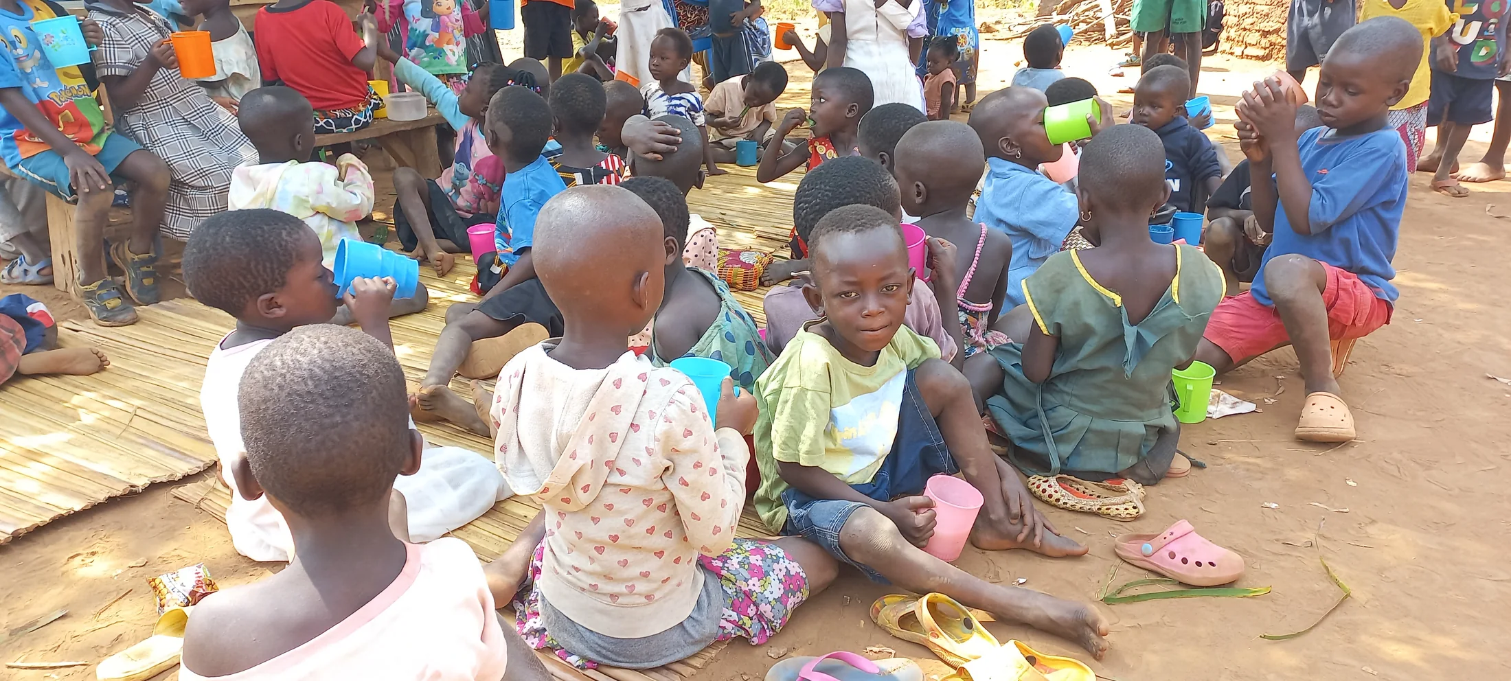 Children sitting together outdoors, many holding cups.